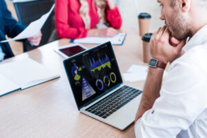 Teilansicht eines nachdenklichen Geschäftsmannes, der mit Papieren und Kollegen am Arbeitsplatz auf Laptop-Bildschirm